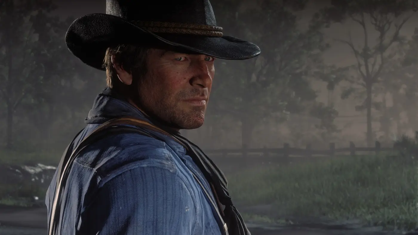 Capture officielle de Red Dead Redemption 2 : gros plan du visage d'Arthur Morgan sous son chapeau noir, barbe naissante, peau marquée et détaillée, devant une forêt brumeuse